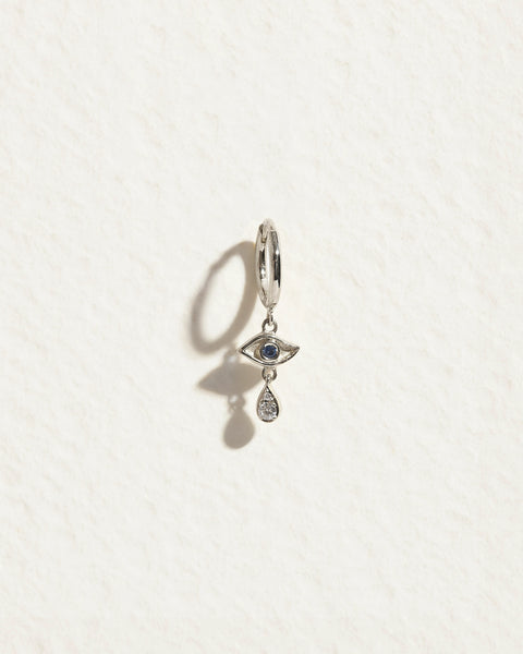 Front view of Crying Eye Huggie earring showcasing a sapphire eye and dangling teardrop-shaped accent