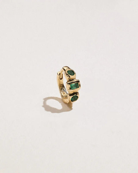 Angled view of a 14K yellow gold piercing huggie showcasing two emeralds framing a baguette emerald in molten bezel settings